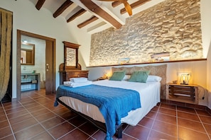 Un dormitorio con pared de piedra, cama con manta azul, mesita de noche de madera y espejo.