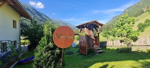 Une cabane en bois, dotée de murs d’escalade et d’une balançoire, se trouve dans une cour verdoyante, avec une montagne en toile de fond.