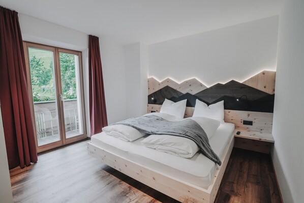 Ein Schlafzimmer mit einem hölzernen Bett, einem Nachttisch, einem Fenster mit Vorhängen und Blick ins Grüne.