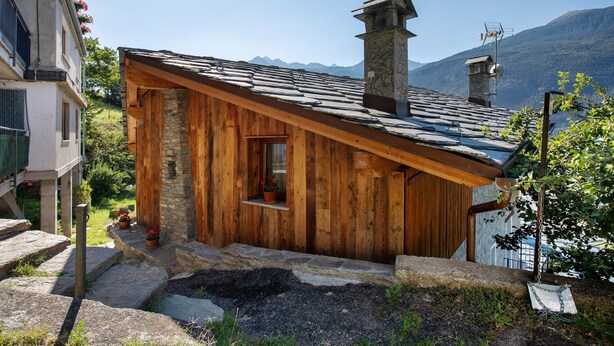 Una cabaña de madera con chimenea de piedra, escalones de piedra y una ventana con vistas a las montañas.