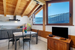 A modern kitchen with a dining area, wooden ceiling beams, a large window with a mountain view, and a television on a wooden cabinet.