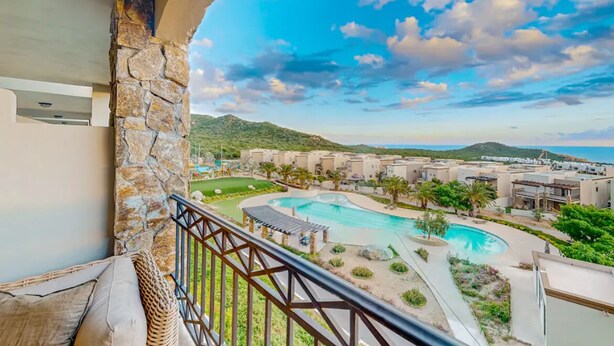Un balcón con pilares de piedra que da a una zona de piscina con vistas a un complejo turístico y colinas al lejano.