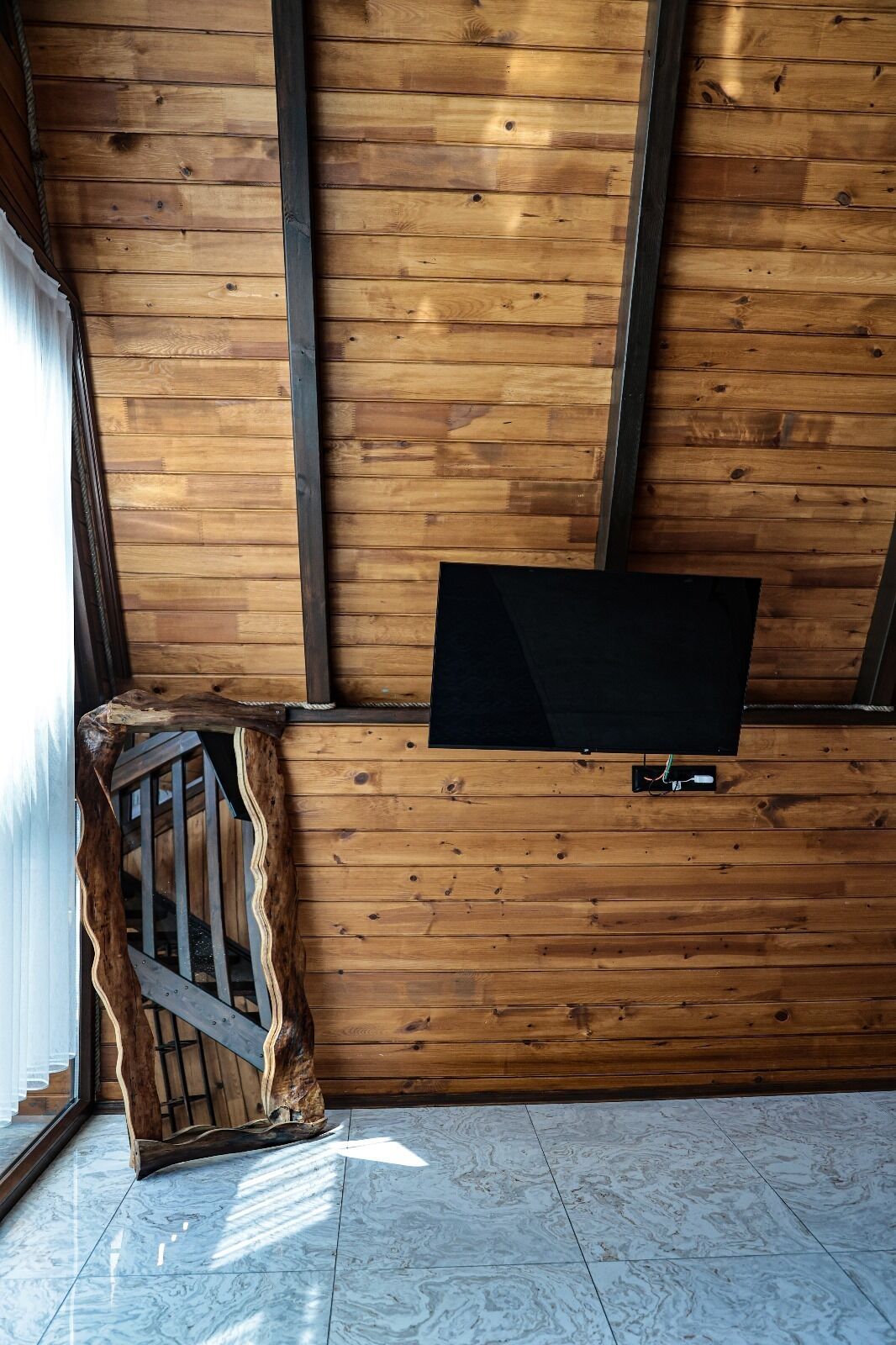 Una habitación con paredes de madera, un televisor de pantalla plana montado, una escalera con barandilla de madera y piso de baldosas.