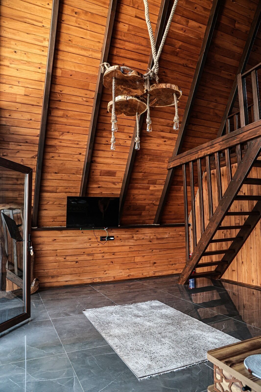 A wooden interior with a sloped ceiling, a mounted TV, a rug, and a wooden bench.