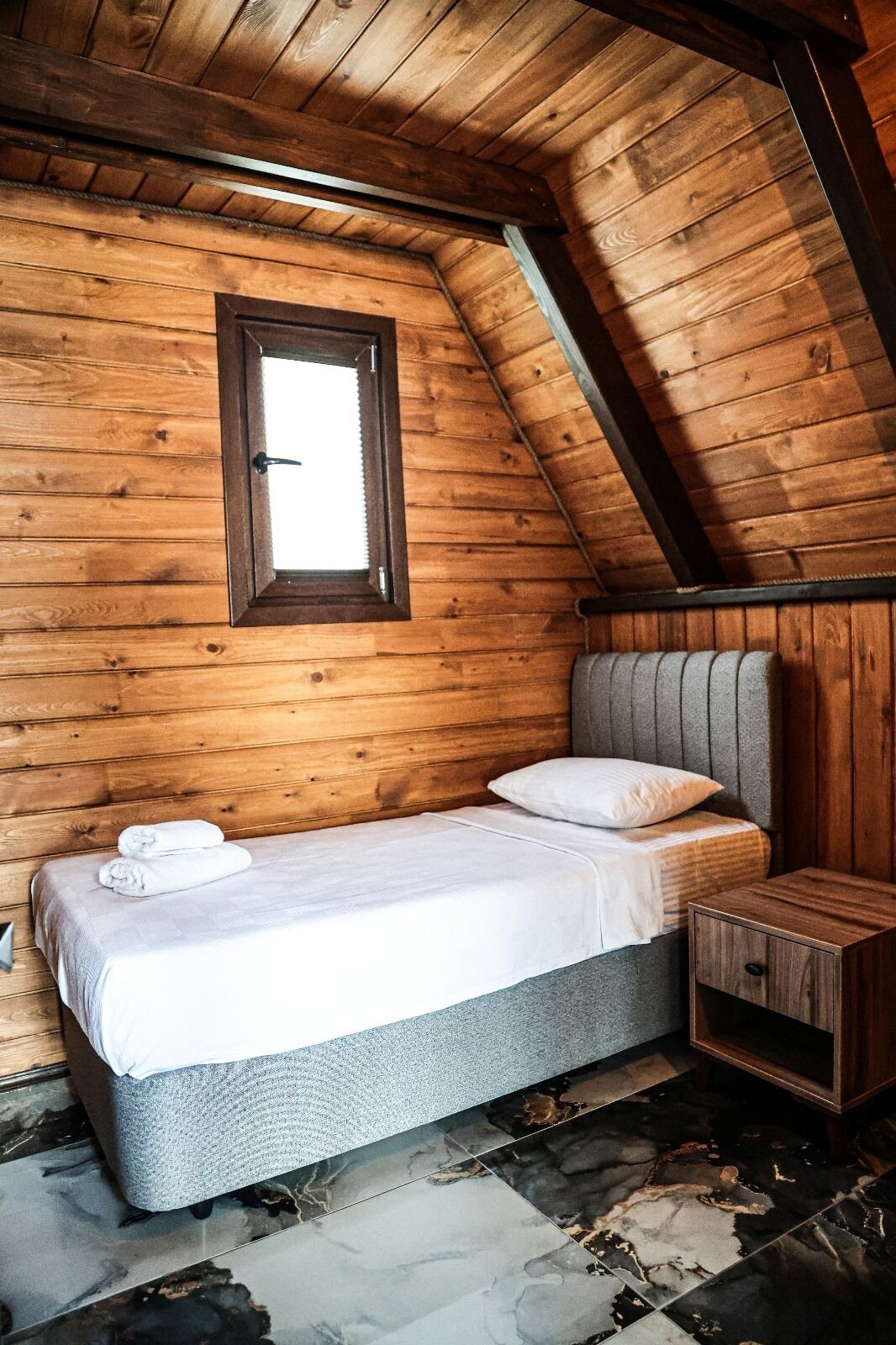 A small, cozy room with wooden walls and a sloped ceiling. It features a single bed with a grey headboard and white bedding, a small wooden nightstand, and a window with a wooden frame.