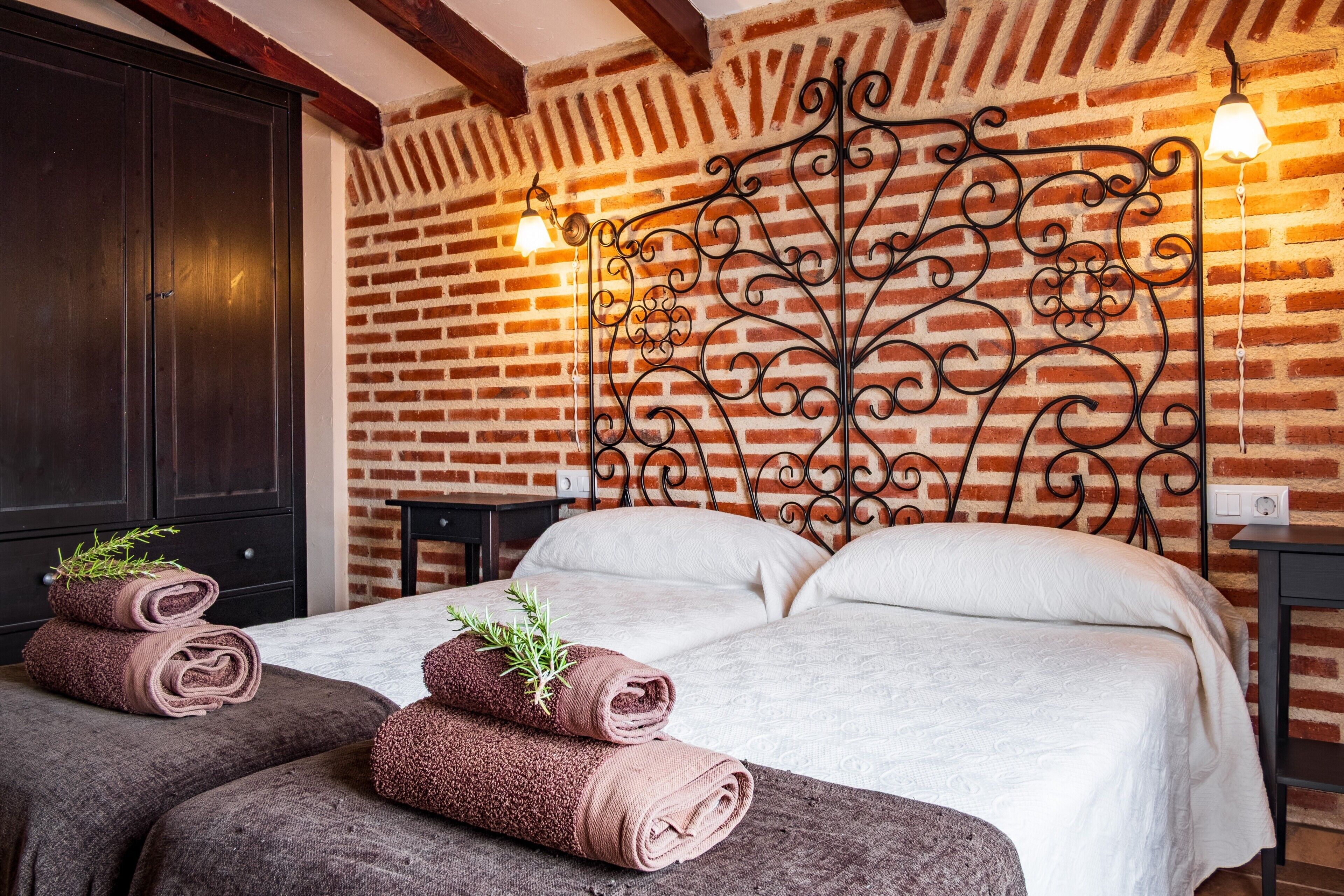 A room with a brick wall, a bed with white bedding, two rolled towels with greenery, and a decorative metal headboard.