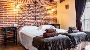 Una habitación con pared de ladrillo, cabecera de hierro forjado, dos camas con ropa blanca y una ventana con cortinas.