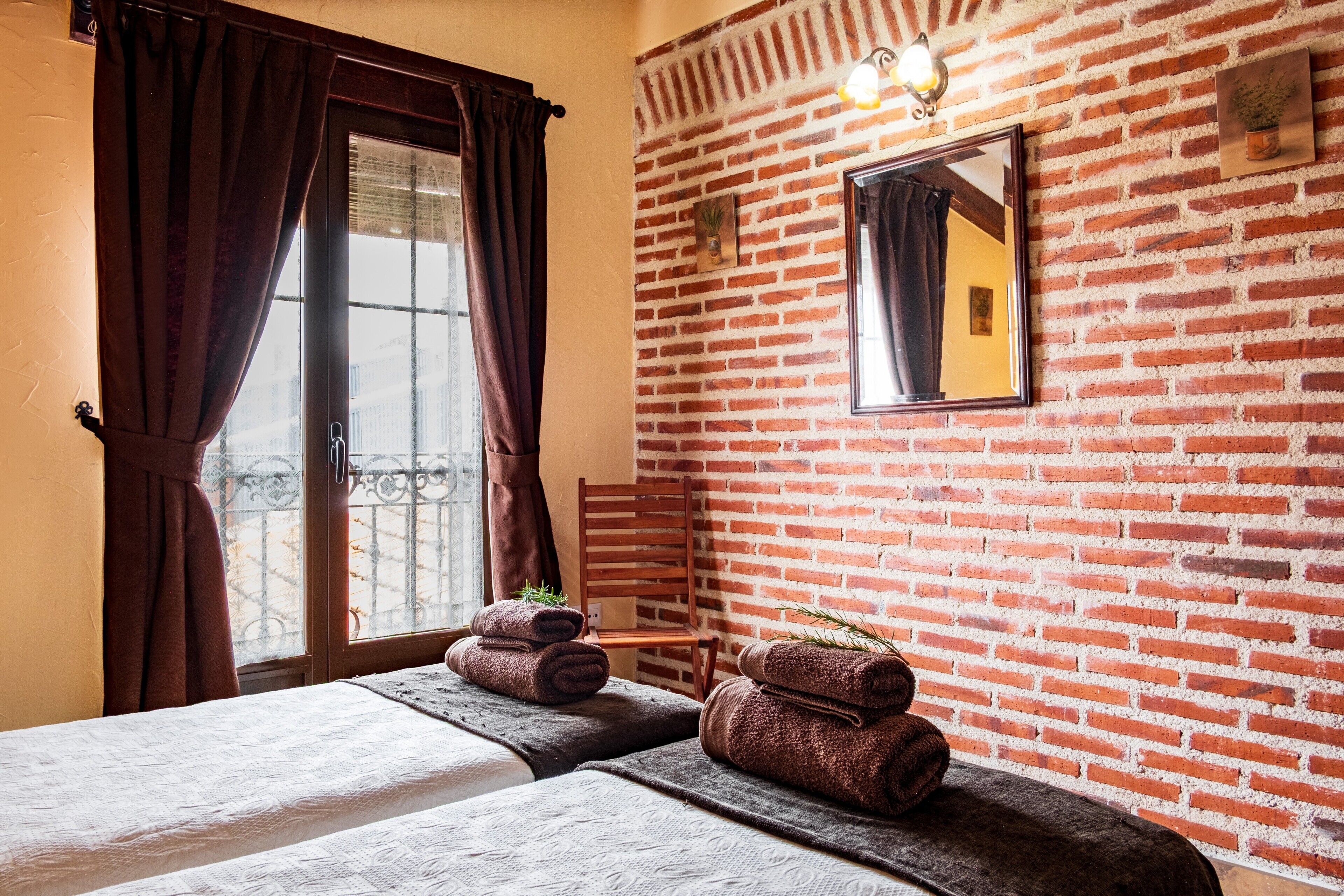 A room with a brick wall, a bed with a white bedspread, a chair, and a window with curtains.