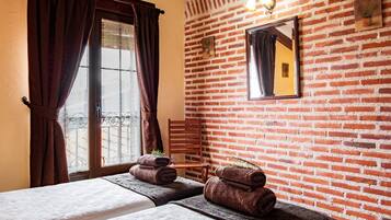 A room with a brick wall, a bed with a white bedspread, a chair, and a window with curtains.