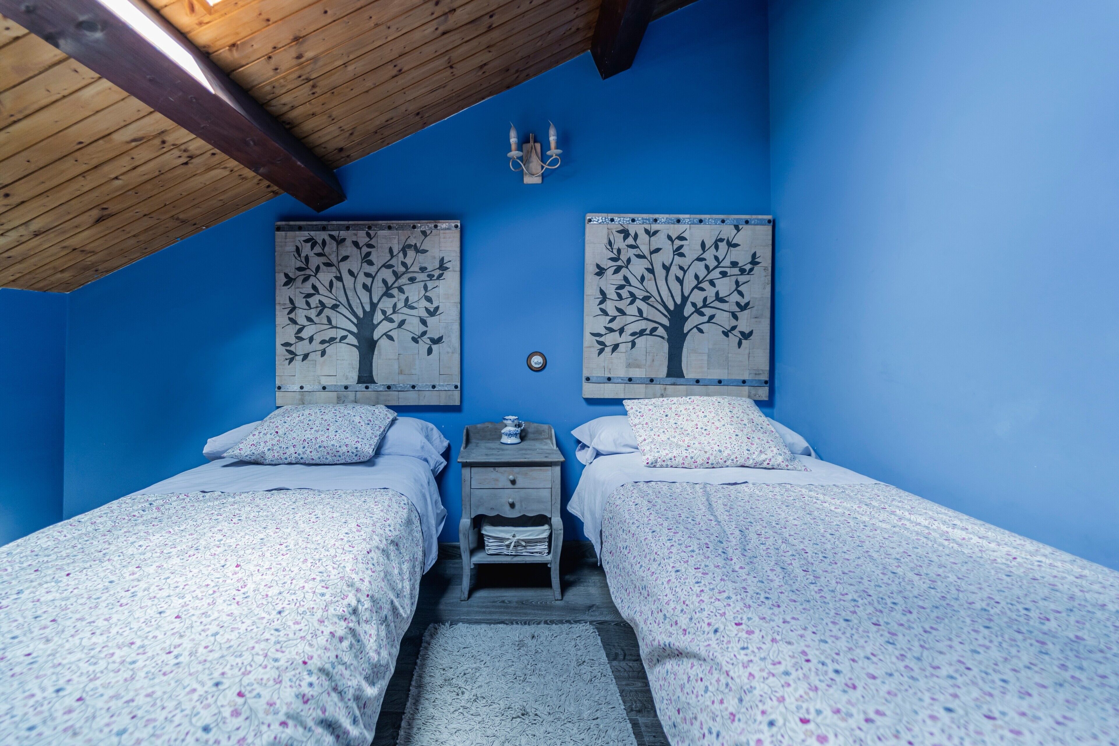 A bedroom with two beds, a nightstand, a wall-mounted lamp, and two tree-themed wall art pieces.