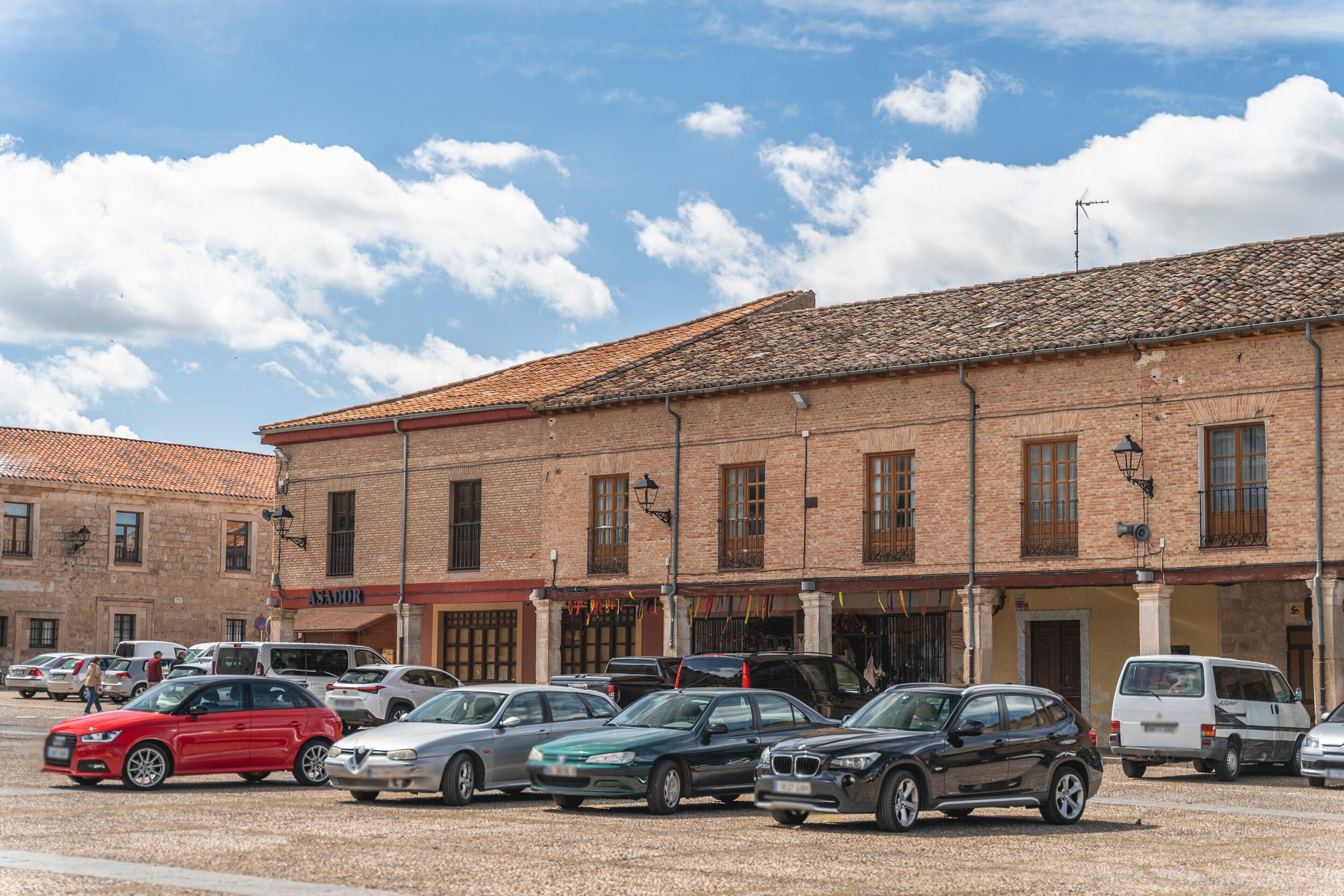 Un estacionamiento con varios autos, un edificio de ladrillo con múltiples ventanas y techo de tejas, y un cielo despejado con nubes.