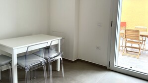 Una stanza con un tavolo bianco e sedie trasparenti, e un'ampia vetrata scorrevole che si apre su una zona esterna con mobili in legno.