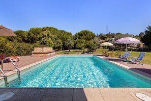 A rectangular swimming pool with clear blue water, surrounded by a stone deck, and shaded by umbrellas.
