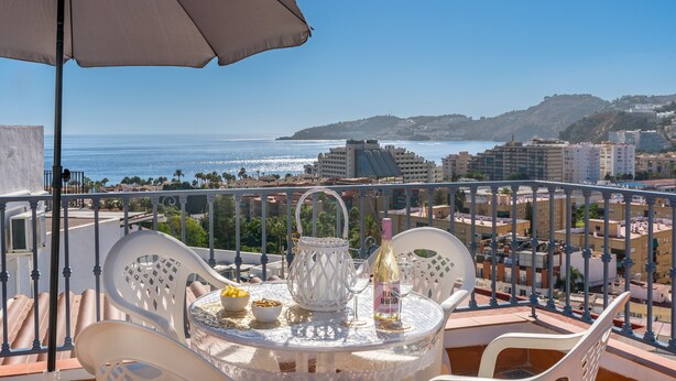 Un balcón con una mesa para dos, una sombrilla blanca y vistas al mar y edificios.