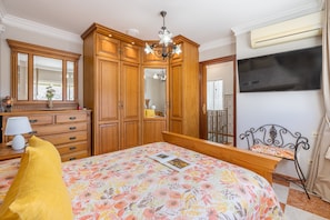 A bedroom with a bed, a dresser, a mirror, a chandelier, a TV, and a balcony.