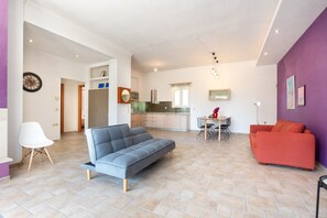 A modern living room with a sofa, a red couch, a dining table with chairs, and a kitchen in the background.