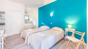 Une chambre d’hôtel avec deux lits, une chaise en bois, une table de chevet avec une lampe, et une salle de bain avec un miroir et un porte-serviettes.