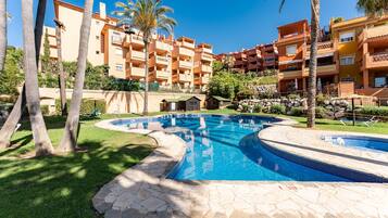 A swimming pool surrounded by stone tiles, palm trees, and multi-story buildings with balconies.