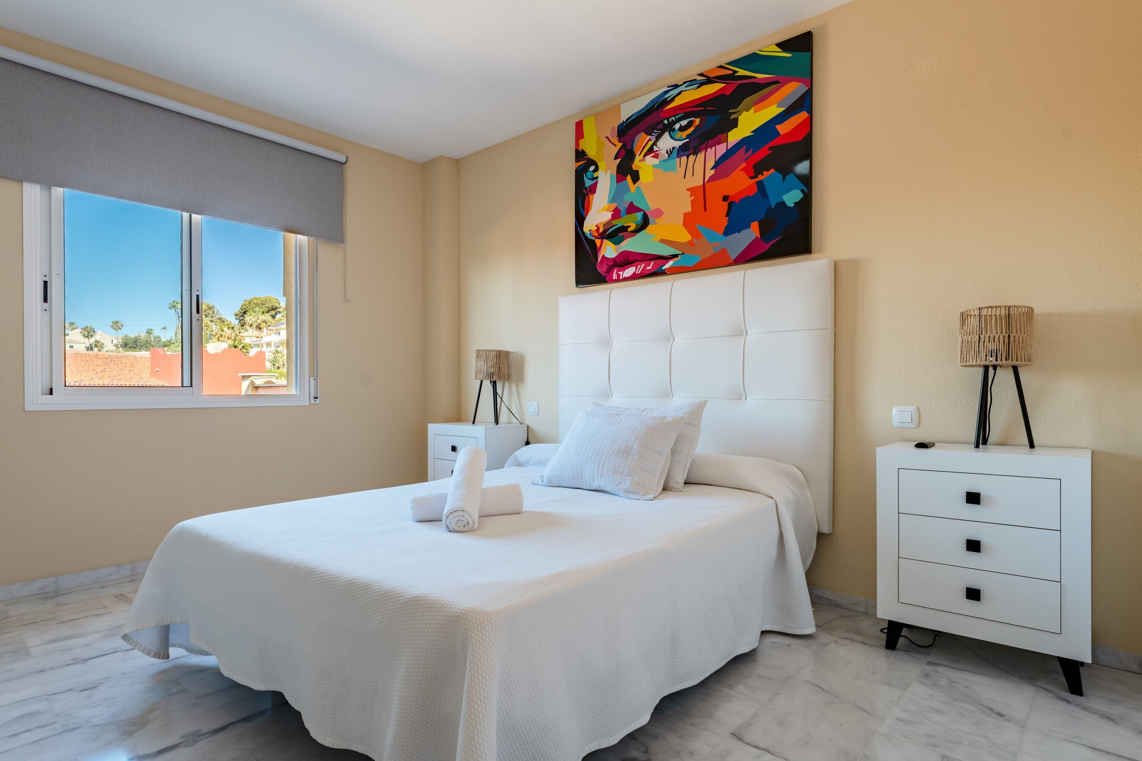 A bedroom with a bed, a nightstand, a lamp, a painting, and a window with a view.