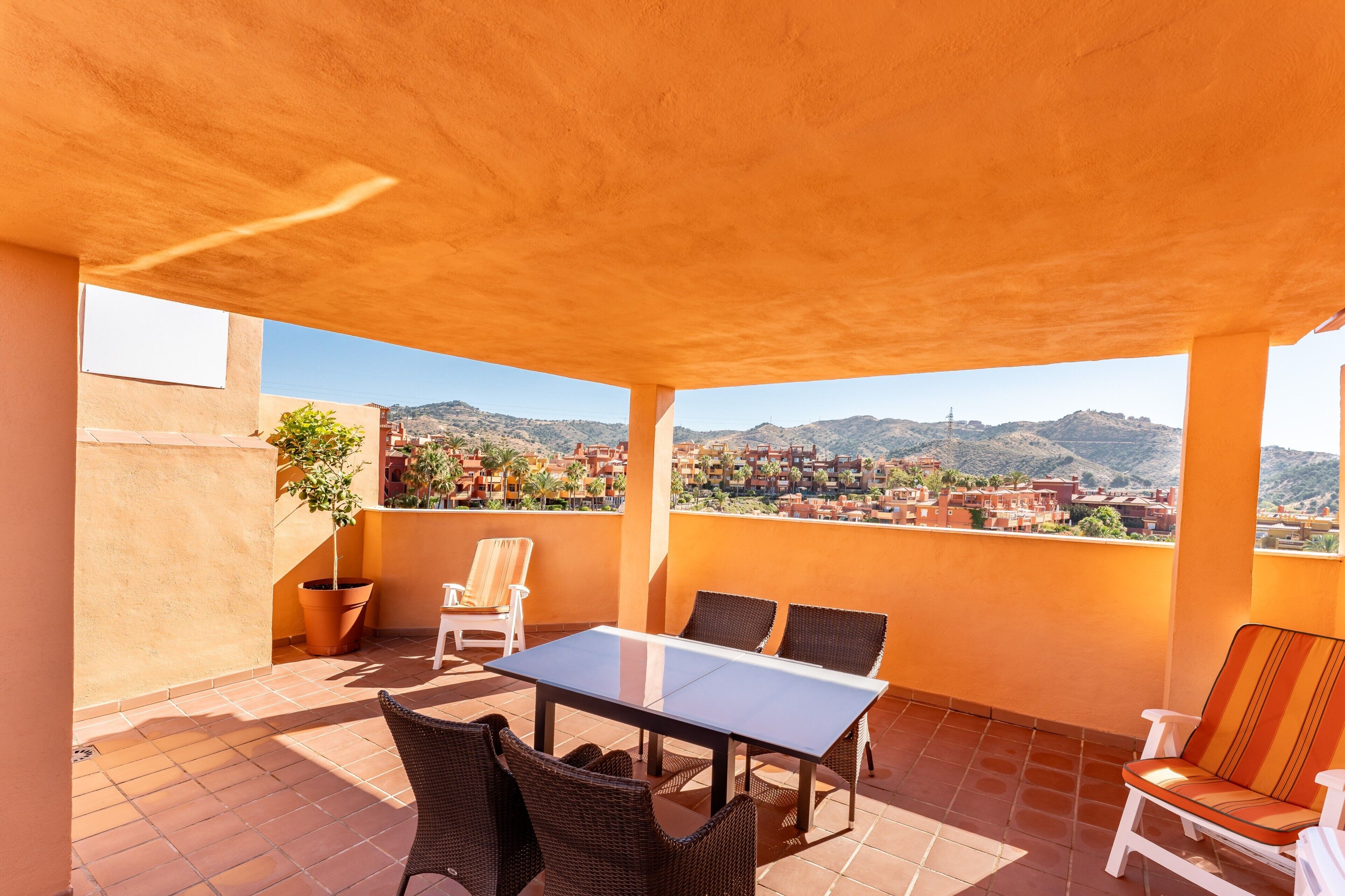 Eine überdachte Terrasse mit Tisch und Stühlen, ein Blumentopf und der Blick auf eine Stadt und Hügel im Hintergrund.