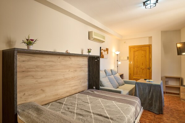 A bedroom with a wooden headboard, a bed with a patterned bedspread, a small table with a lamp, and a wooden wardrobe.