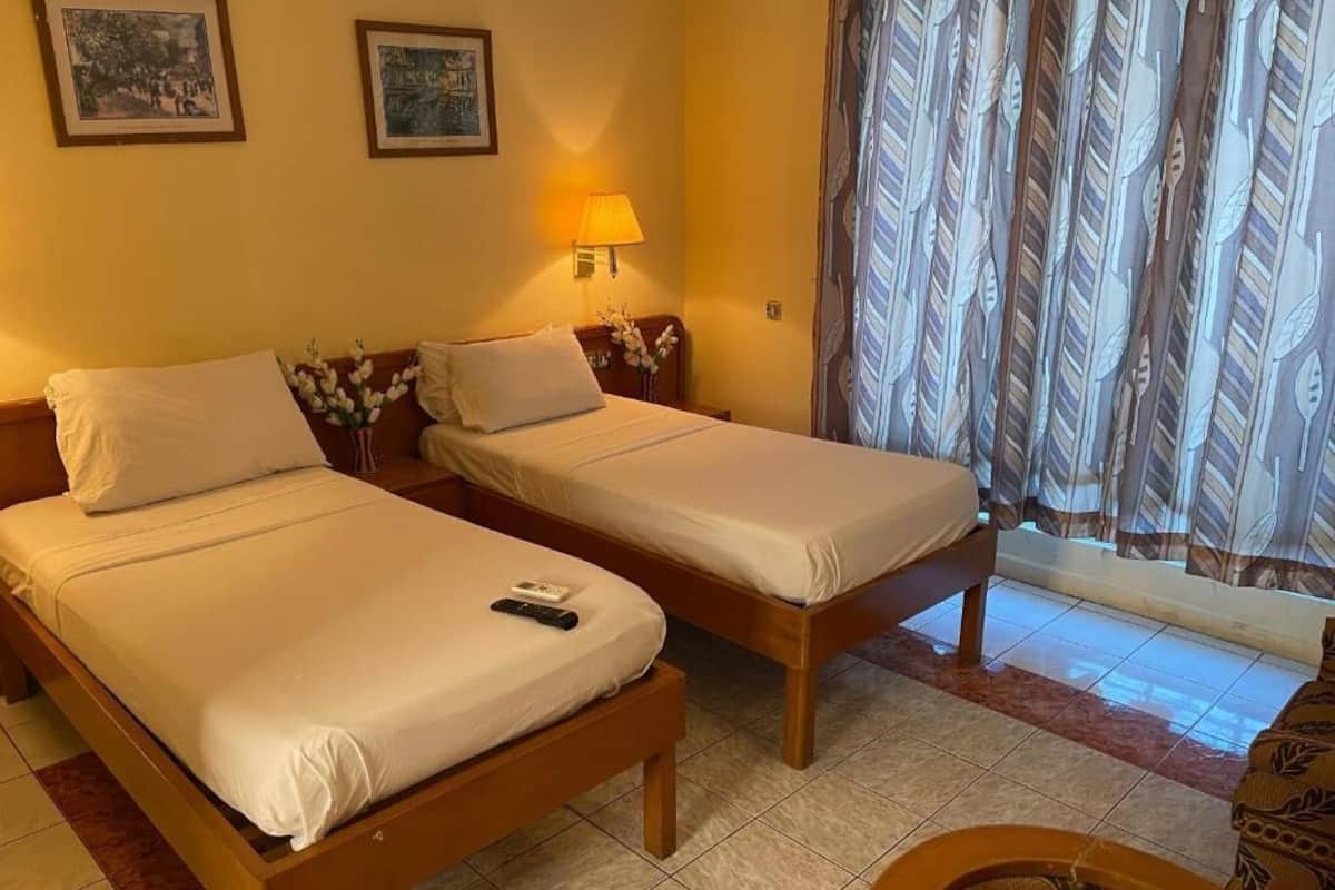 A hotel room with two single beds, a nightstand with a lamp, a small table with a vase of flowers, and a window with patterned curtains.
