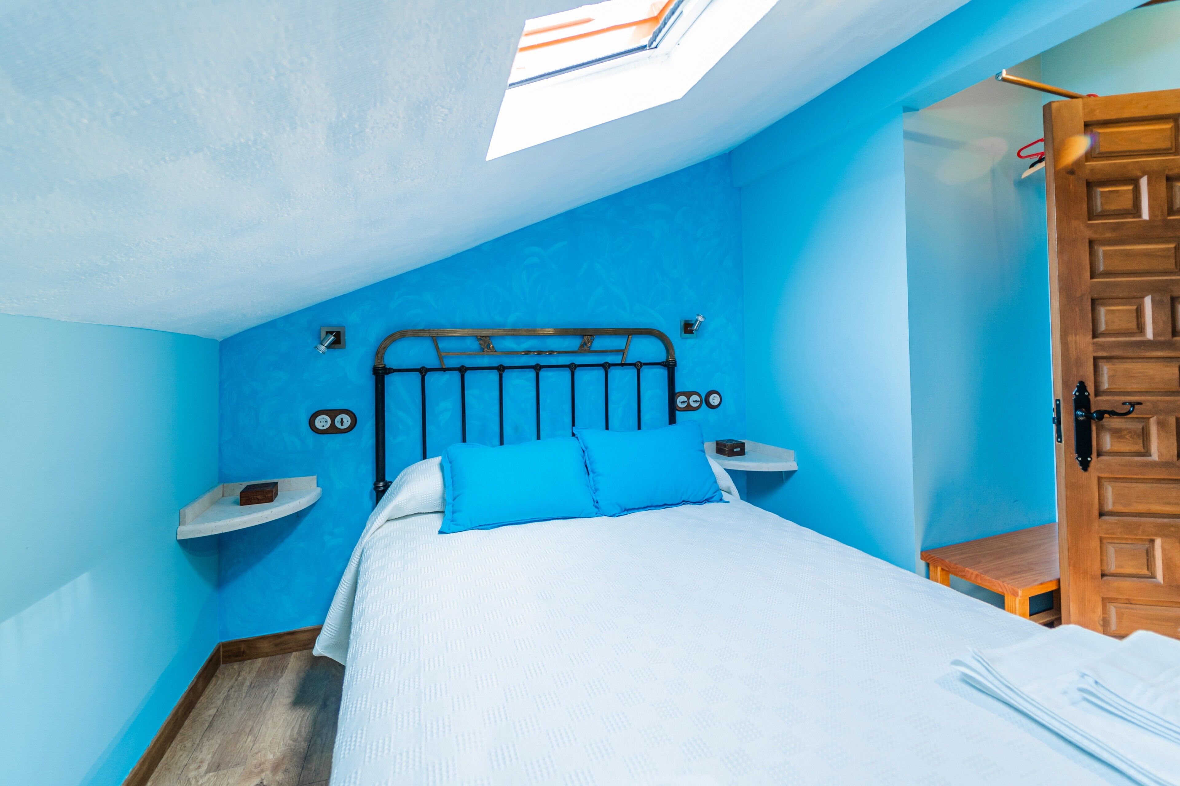 A bedroom with a skylight, a bed with blue bedding, a wooden door, and a small bedside table.