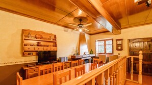 Un coin repas en bois avec une longue table et des chaises, un ventilateur de plafond, et un espace cuisine avec des armoires en bois et une cuisinière.