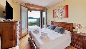 A bedroom with a bed, a TV, a wooden dresser, a nightstand, and a view of the outdoors. - Happy Villa Apartment with Lake View, Private Terrace and Wi-Fi (San Felice del Benaco)