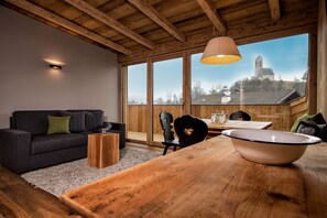 A modern living room with a wooden ceiling, a sofa, a dining table, and a view of a church through large windows.
