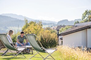 Two people sitting on lawn chairs in a grassy area with a house and hills in the background.