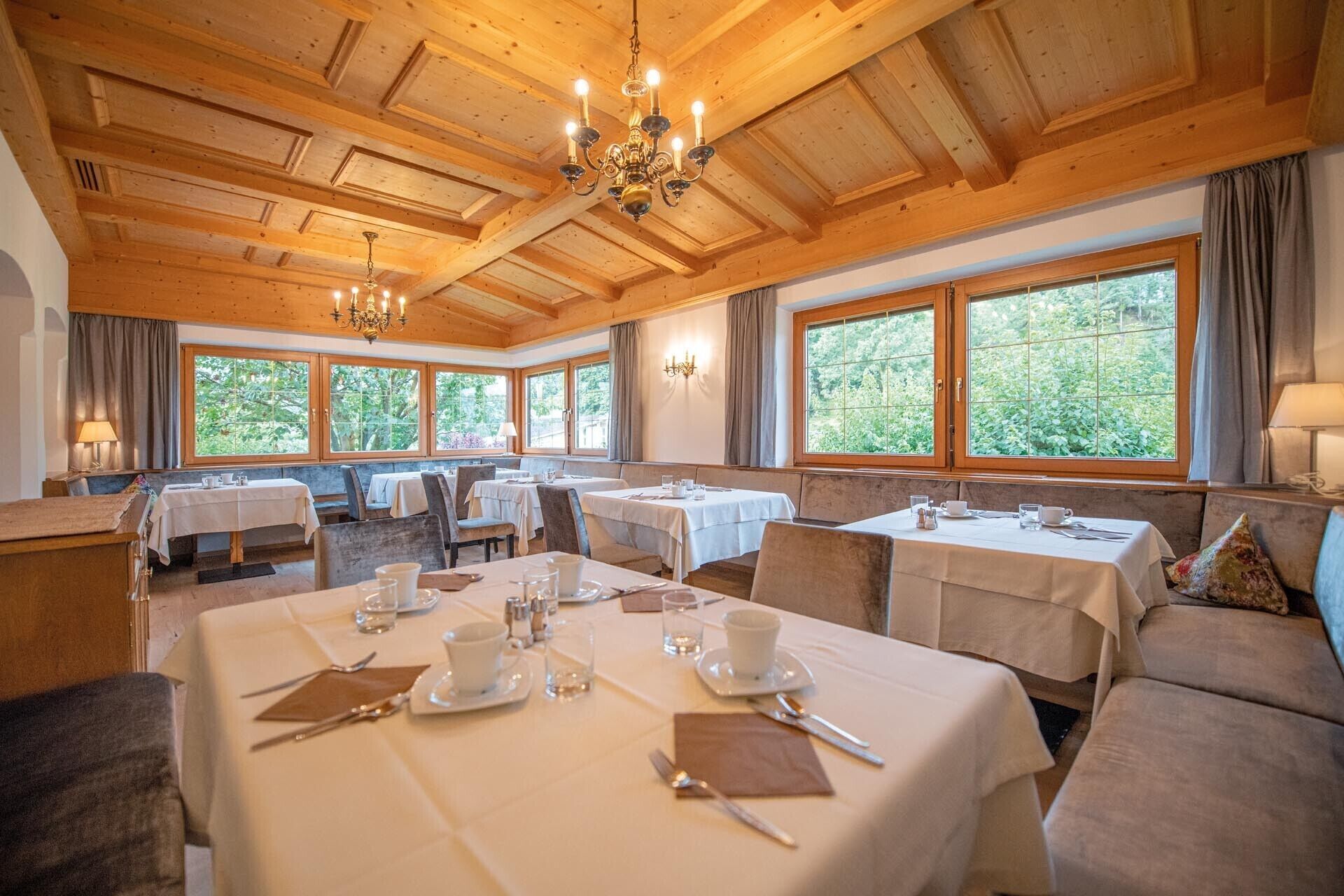Zona pranzo con tavoli apparecchiati, ampie finestre con vista sul verde e soffitto in legno con lampadari.