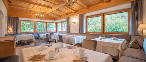 Zona pranzo con tavoli apparecchiati, ampie finestre con vista sul verde e soffitto in legno con lampadari.