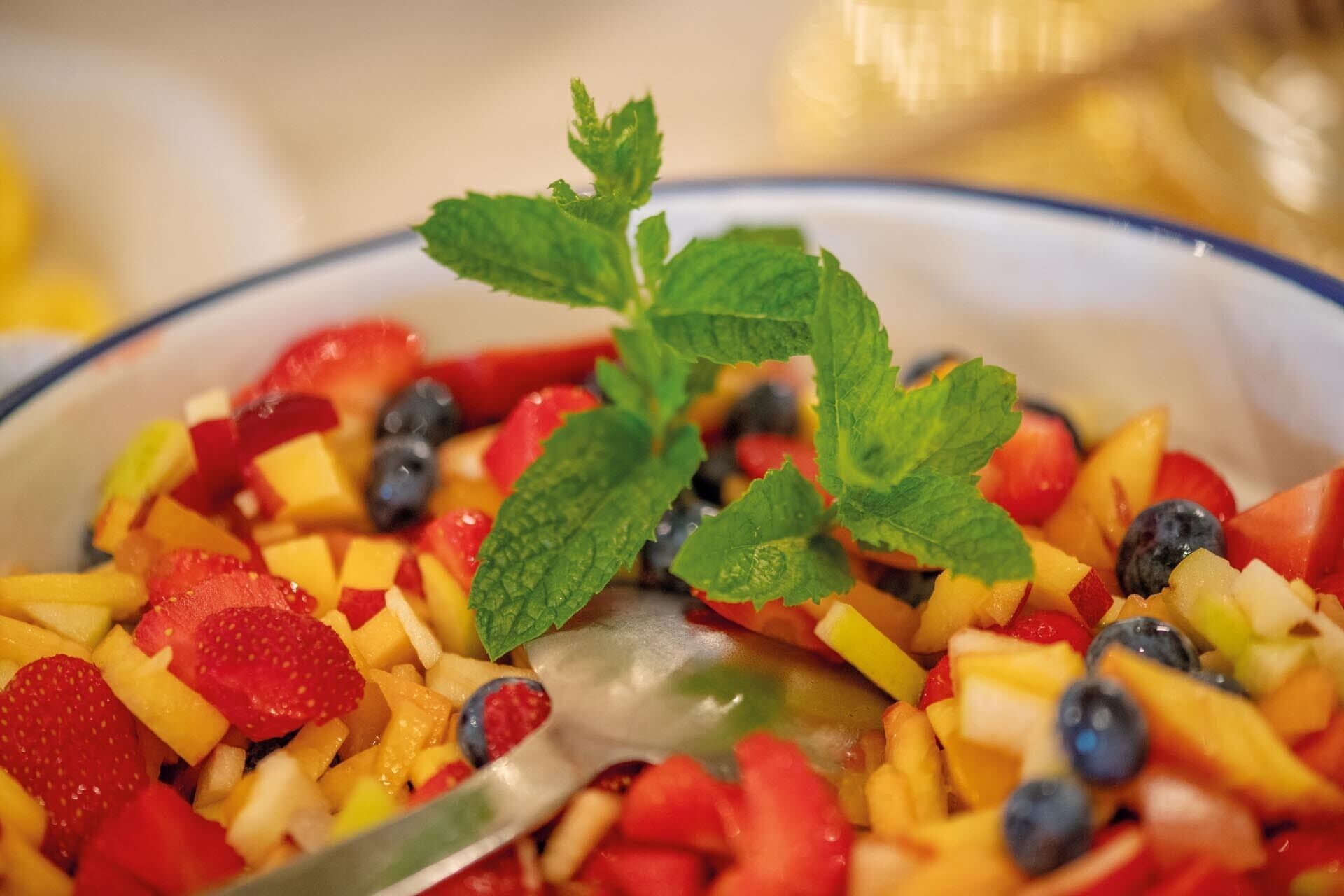 A bowl of fruit salad with strawberries, blueberries, and mango chunks, garnished with mint leaves.