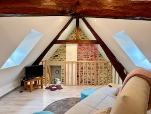 A loft bedroom with a sloped ceiling, wooden beams, a television, a sofa, and a small table.