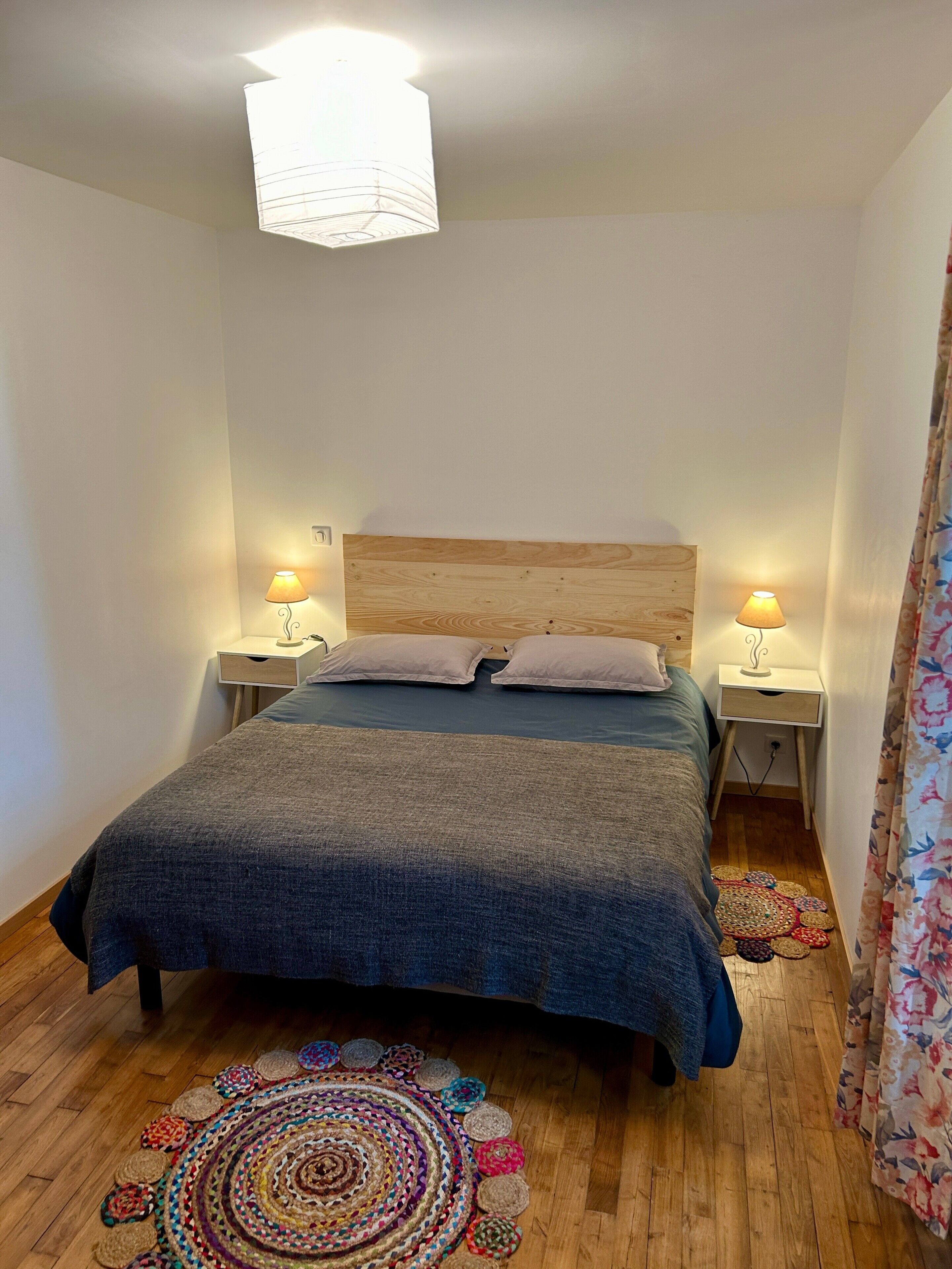 Ein Schlafzimmer mit einem hölzernen Kopfteil, zwei Nachttischen mit Lampen, einem Bett mit blauer Decke und einem farbenfrohen runden Teppich auf dem Boden.