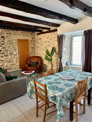 Uma sala de estar aconchegante com parede de pedra, porta de madeira, poltrona de couro, mesa de jantar com toalha de mesa estampada em folhas e um vaso com planta.