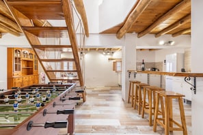 Una habitación amplia con una mesa de fútbol, taburetes de madera y una zona de cocina.