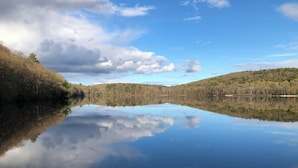 Un lac paisible aux eaux claires, où se reflètent les arbres et le ciel, entouré de collines et surplombé d’un ciel partiellement nuageux.