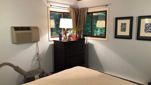 Une chambre à coucher comprenant un lit, une commode, une fenêtre avec des rideaux, un climatiseur et des œuvres d’art encadrées accrochées au mur.