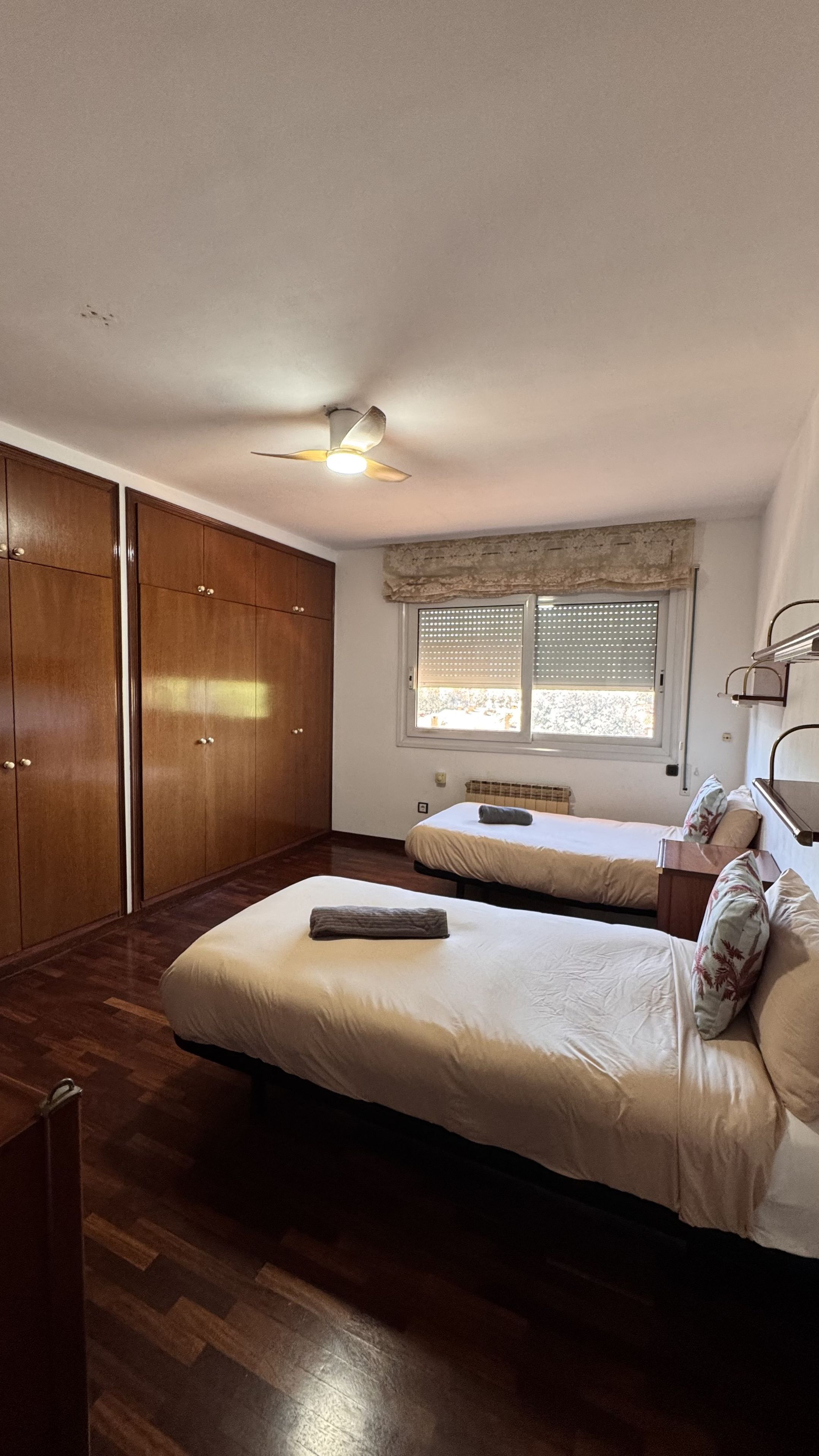 Een slaapkamer met twee bedden, houten kasten, een plafondventilator, een raam met zonwering en een radiator.