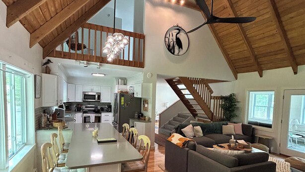 Un vaste espace de vie aménagé dans un style « loft », avec un plafond haut, un coin repas doté de chaises en bois, un canapé gris et un escalier menant à un niveau supérieur.
