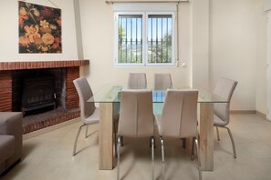 Un comedor con una mesa de vidrio, cuatro sillas, chimenea de ladrillo y ventana con vistas.