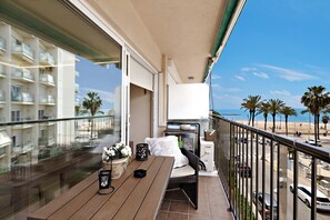 A balcony with a wooden table, a wicker chair, a potted plant, and a view of the beach and palm trees.