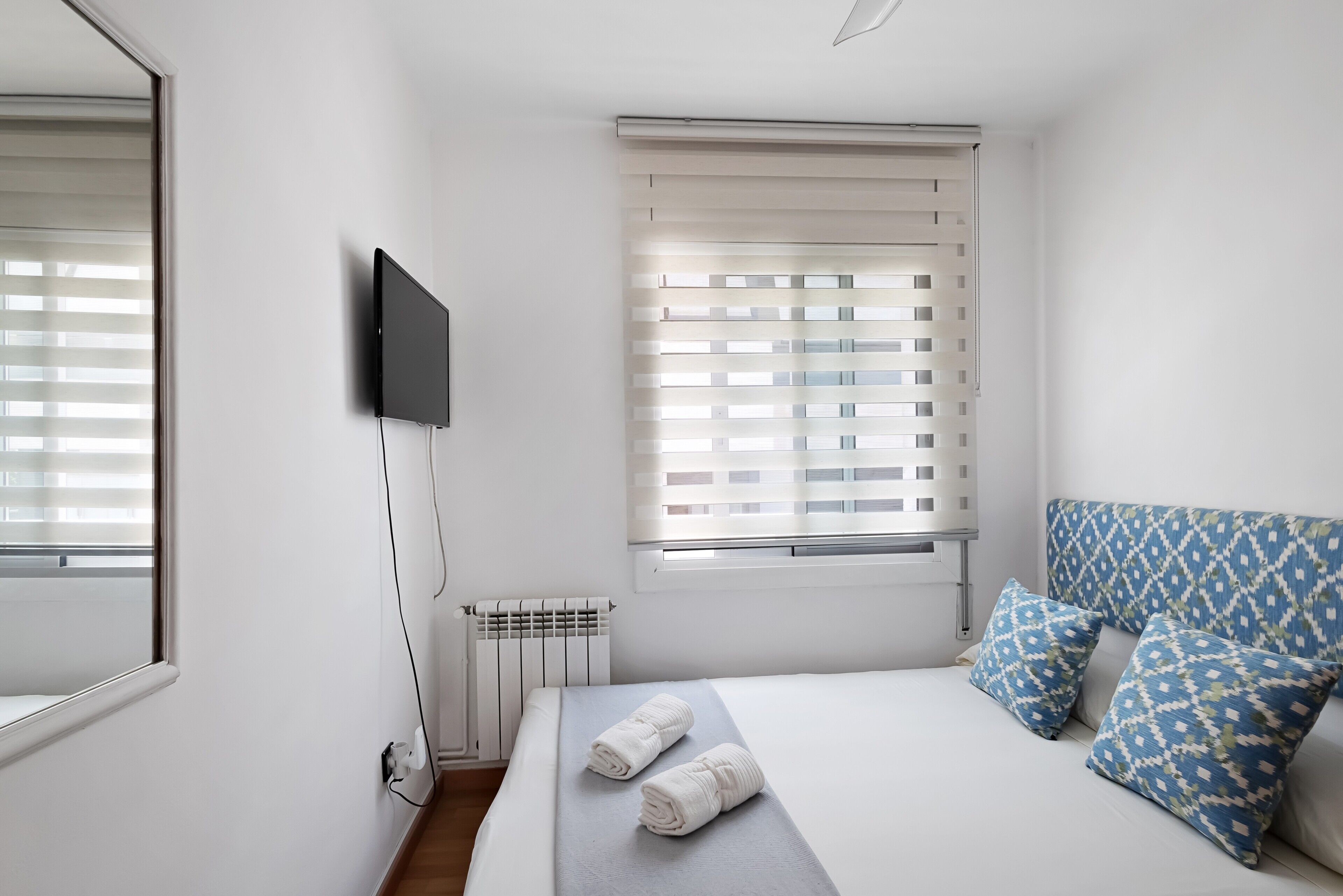 Un dormitorio con cama, dos almohadas, televisor, ventana con persianas y radiador.