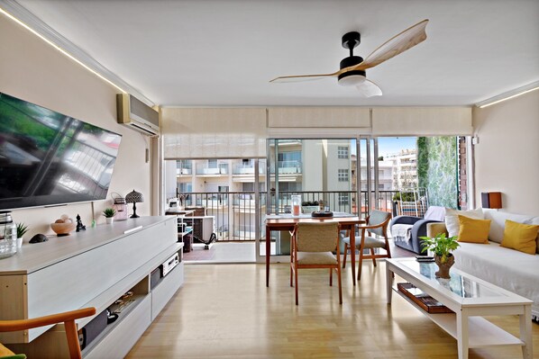 Un salon moderne doté d’un téléviseur à écran plat, d’un ventilateur de plafond, d’un coin repas avec une table et des chaises, et d’un balcon offrant une vue sur les immeubles.