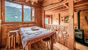 A wooden cabin interior with a dining table, chairs, a stove, and large windows overlooking a forest.