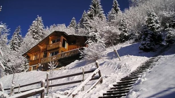 A wooden cabin with a balcony, surrounded by snow-covered trees and a wooden staircase.