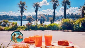 A table with food and drinks, palm trees, and a scenic view of a beach and cityscape. - Brittany Dream in Perros-Guirec – Sea View, Private Terrace, Ideal for Remote Work – High-Speed Wi-Fi (Perros-Guirec)