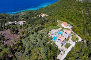 An aerial view of a coastal property with multiple buildings, a swimming pool, and a forested hillside.